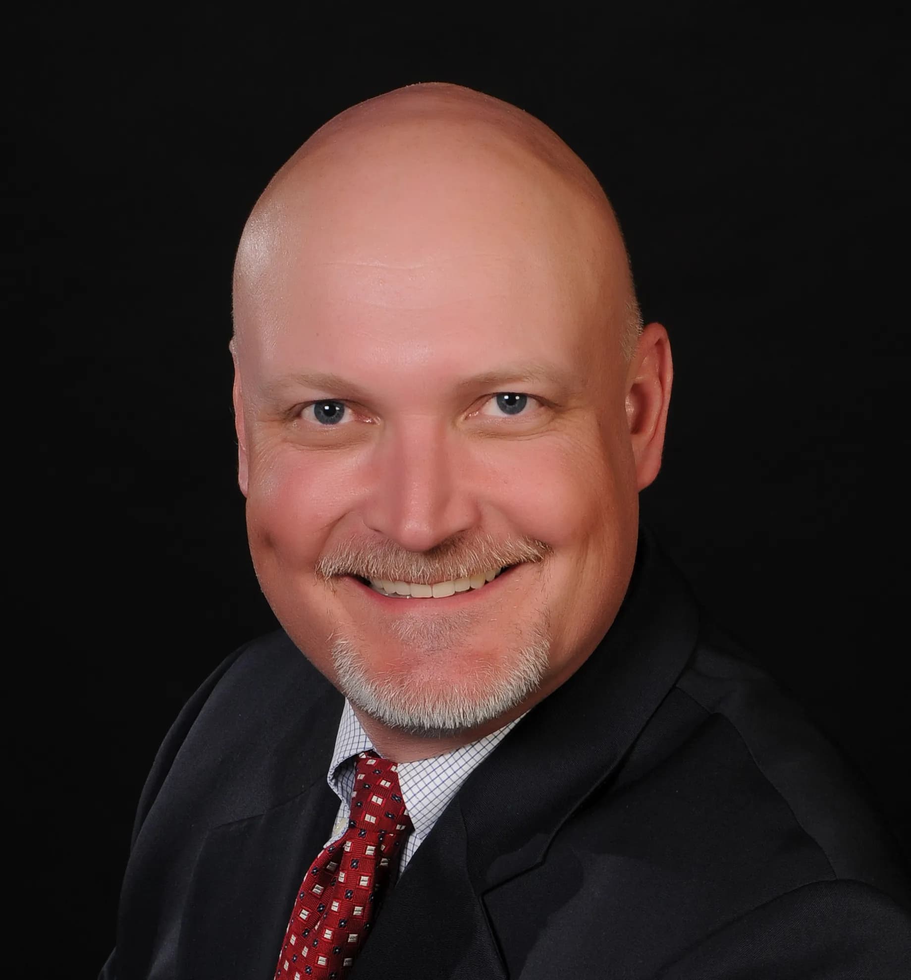Collin Mooney smiling in a black blazer and red tie against a black office background.