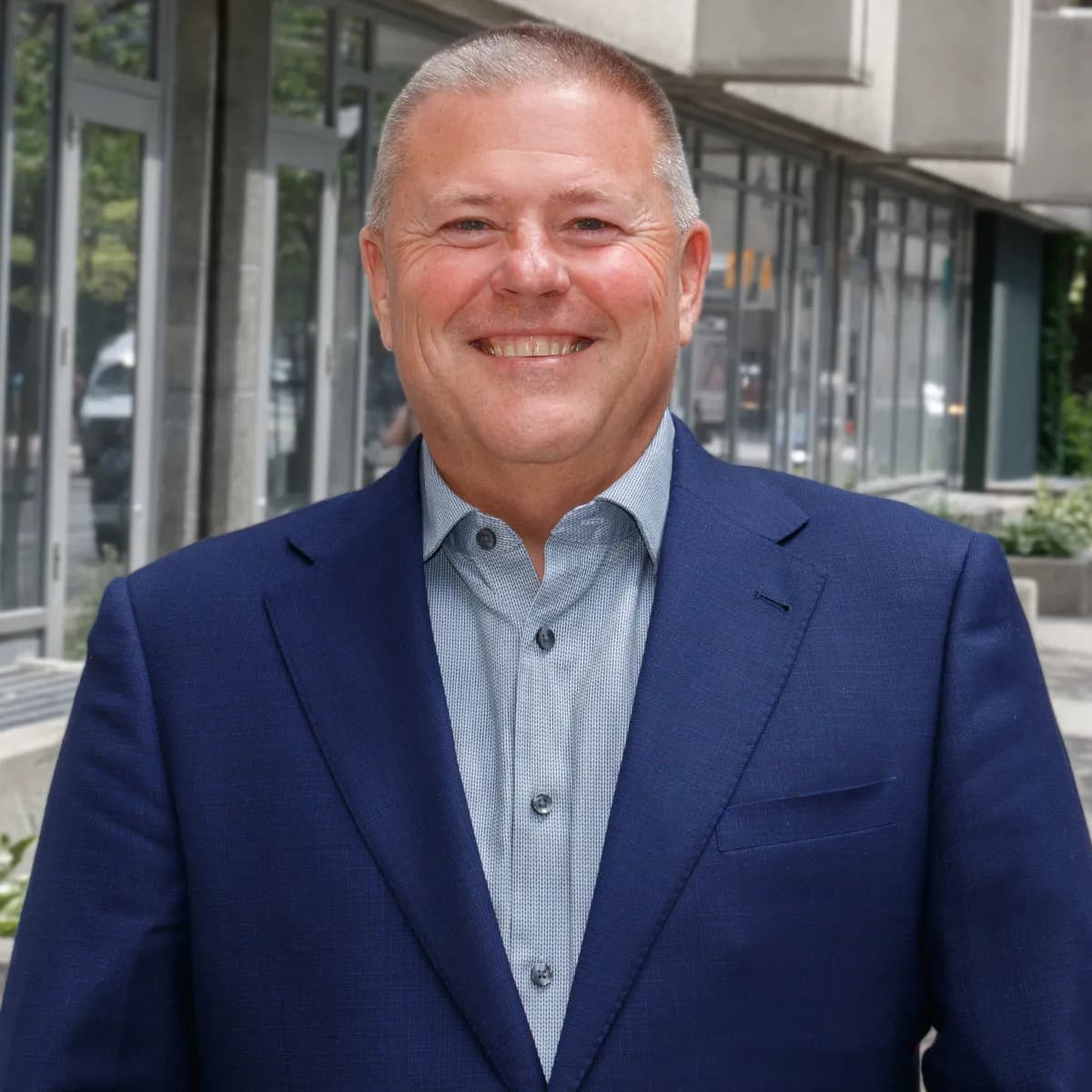 John Sova smiling in a blue blazer against an outdoor urban background.