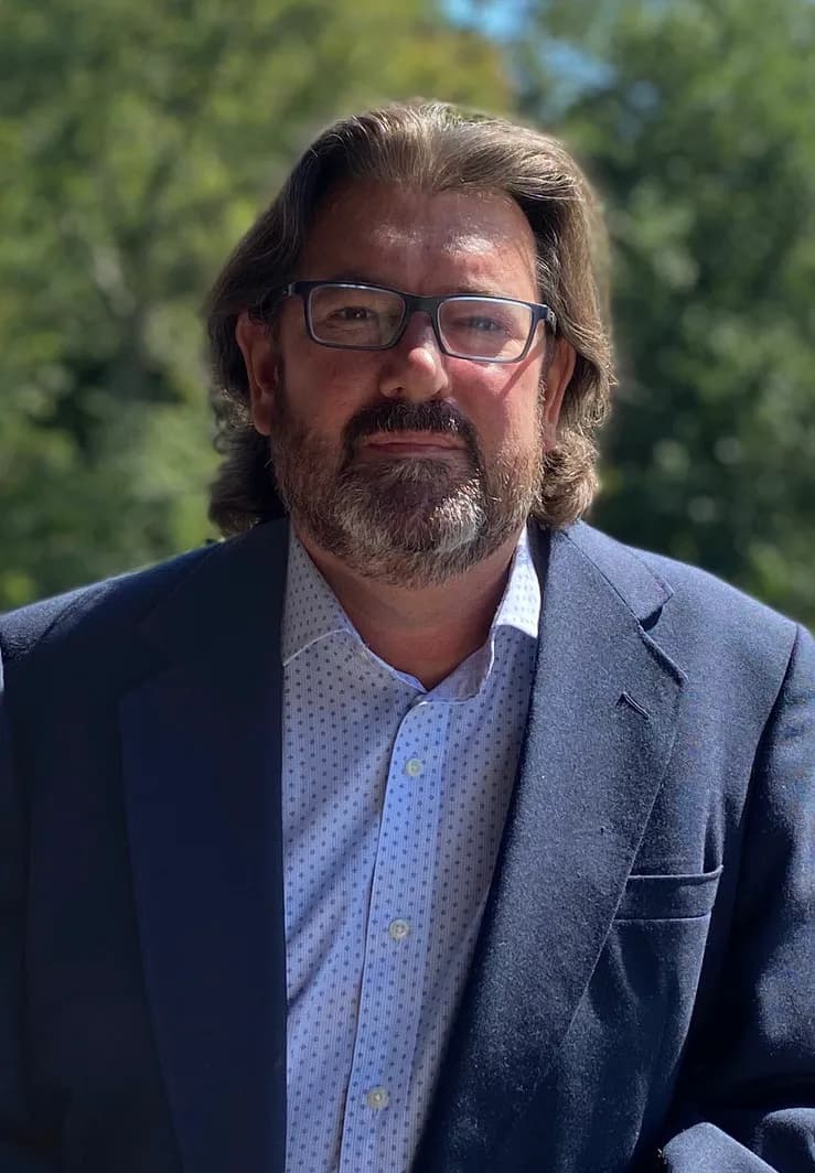 Ken Albrecht in a blue blazer against an outdoor treeline background.