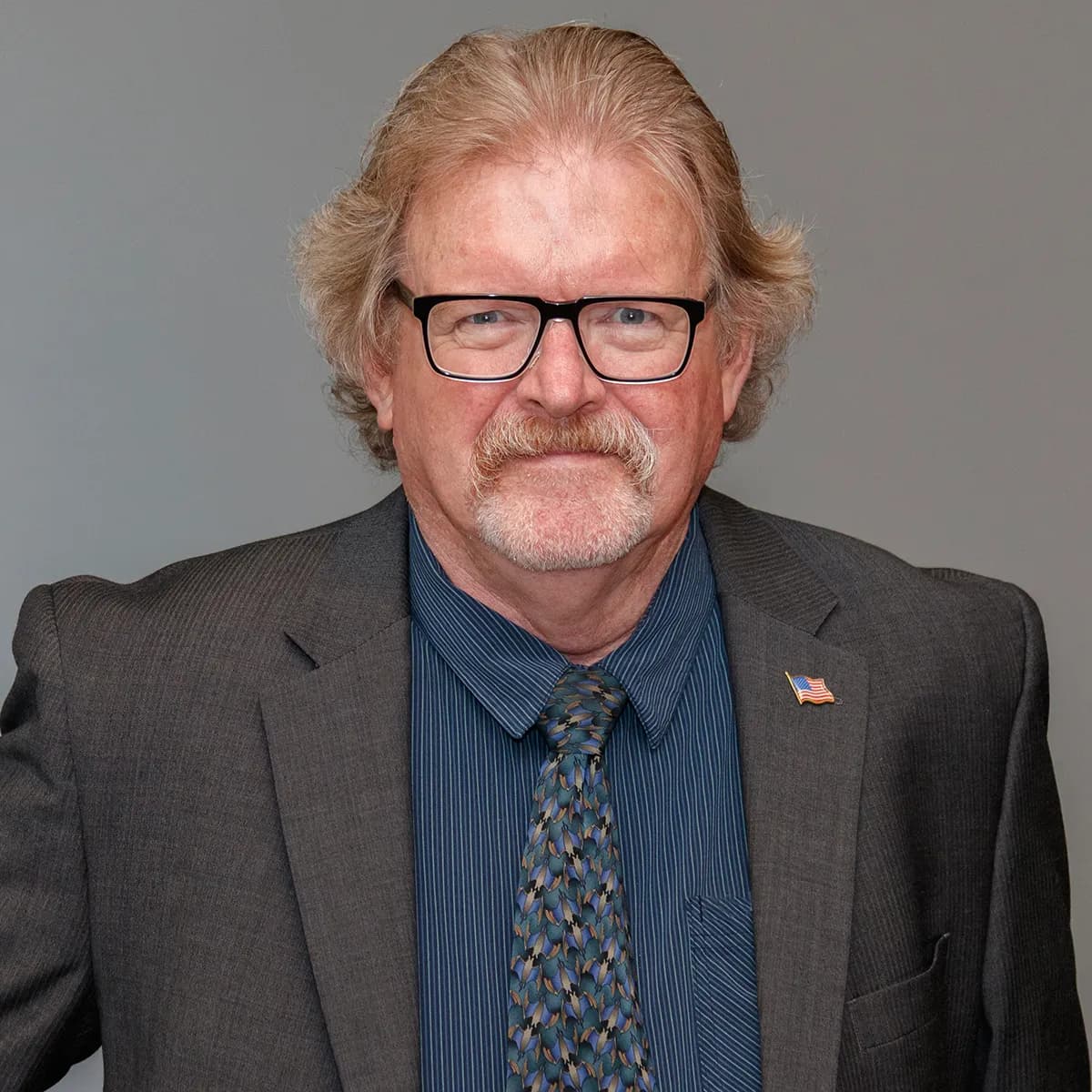 Mark Mills smiling in a grey blazer and blue tie against a gray office background.
