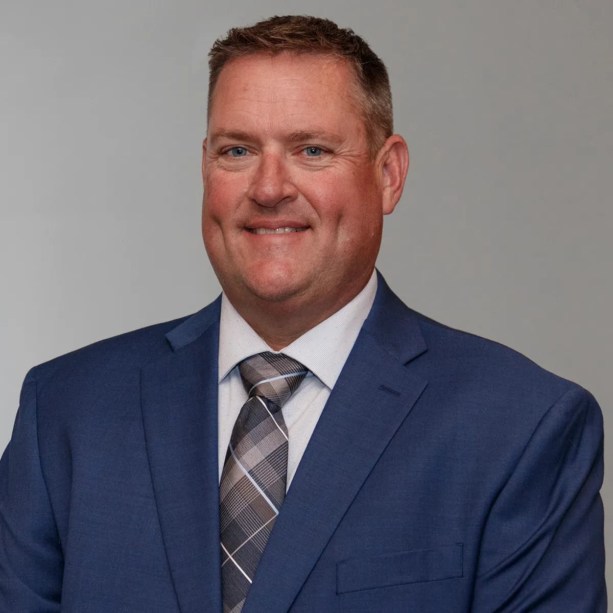 Matt Mullen smiling in a blue blazer and grey tie against a gray office background.