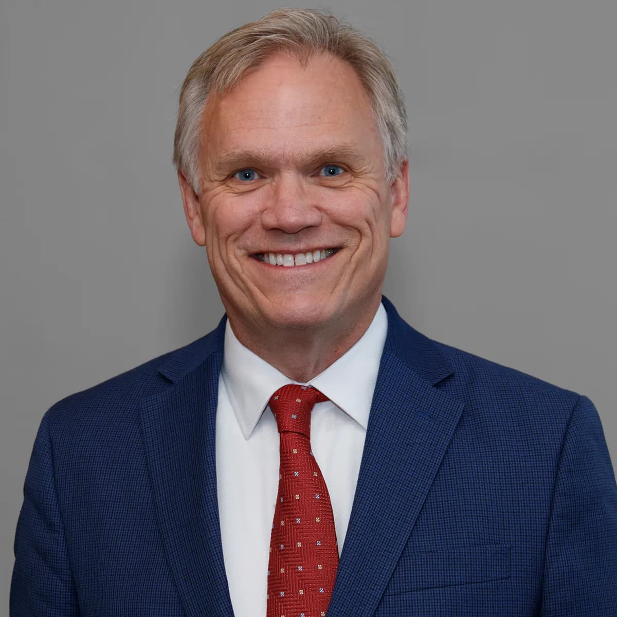 Tome Kean smiling in a blue blazer and red tie against a gray office background.